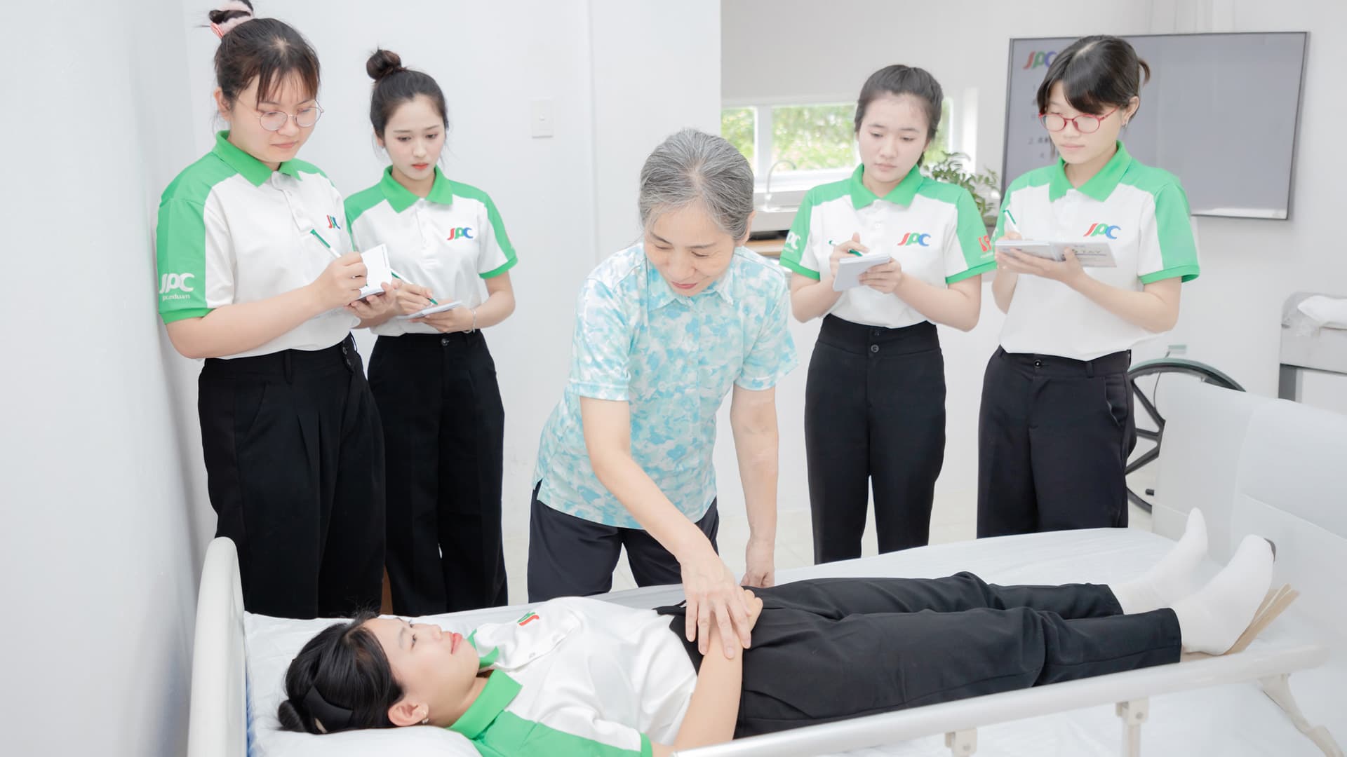Giảng viên hướng dẫn thao tác chăm sóc trên giường bệnh, học viên mặc đồng phục JPC đứng ghi chép